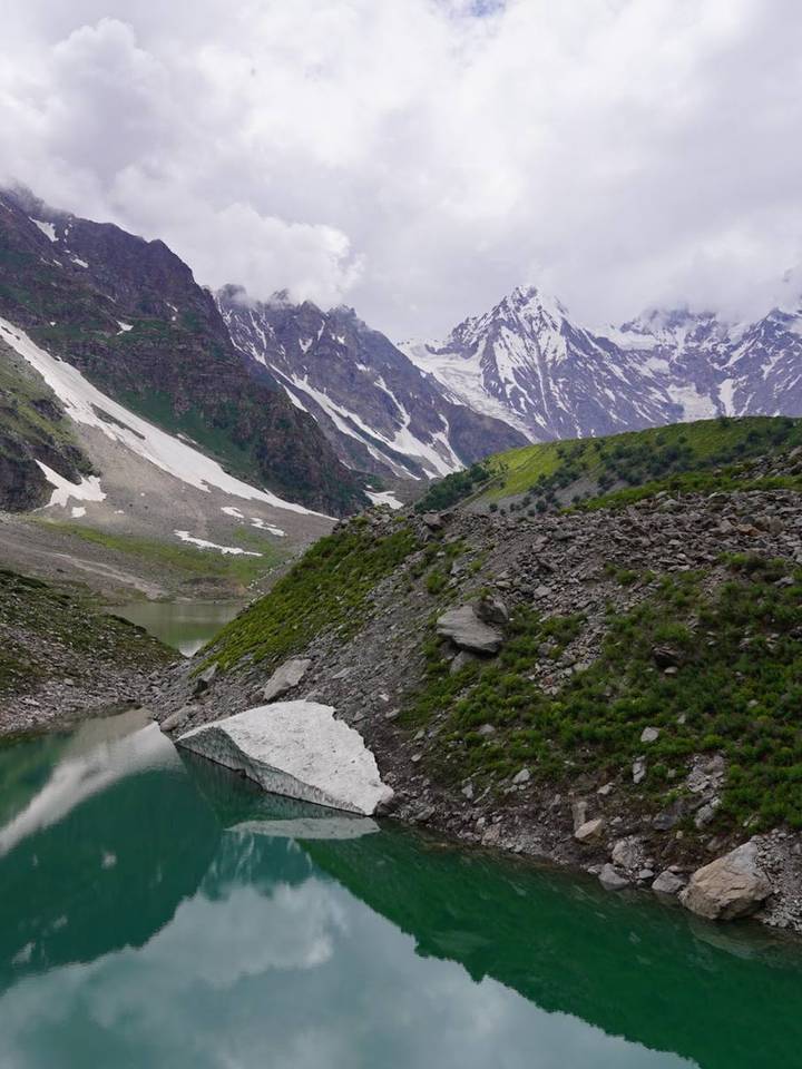 A green glacial lake with snow banks below clouded peaks near Skardu