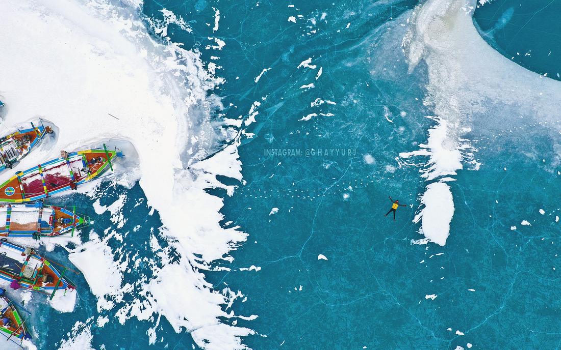 Drone view of a traveller lying on a frozen turquoise lake beside painted boats