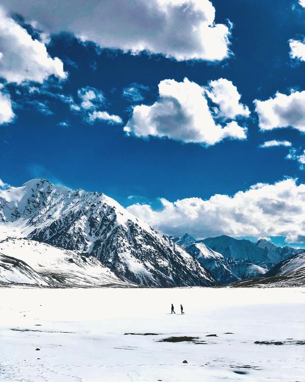 Two hikers crossing a snow plain below peaks and big clouds